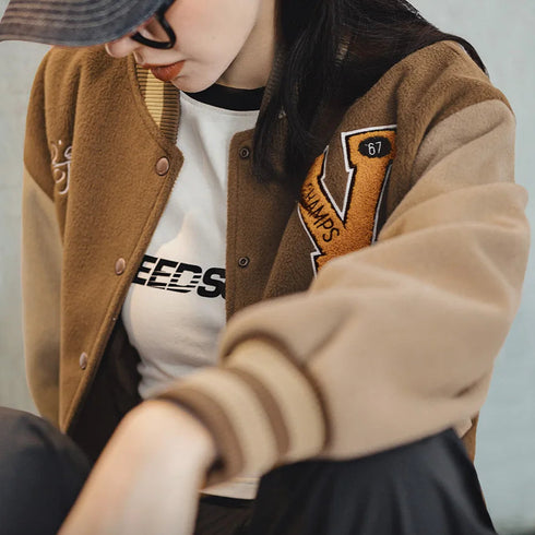 Person wearing a brown letterman jacket with a logo, sitting against a neutral background.